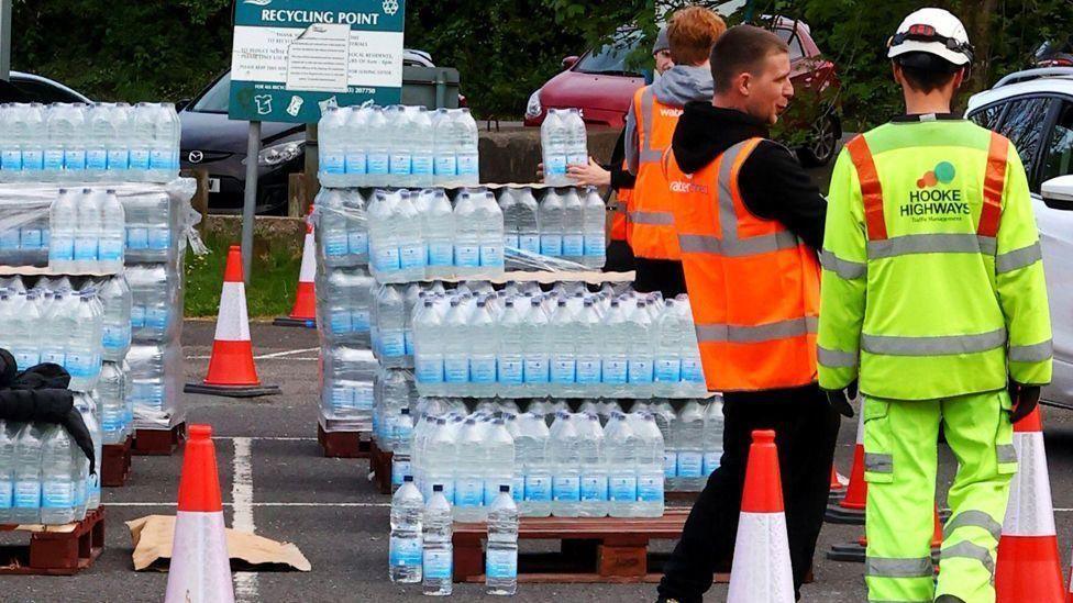 Several pallets of water bottles with road cones and a number of people in hi-vis outfits.