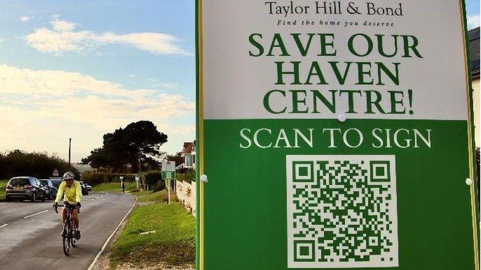 A sign next to a road reads: "Save our haven centre!" It invites people to sign a printed QR code.