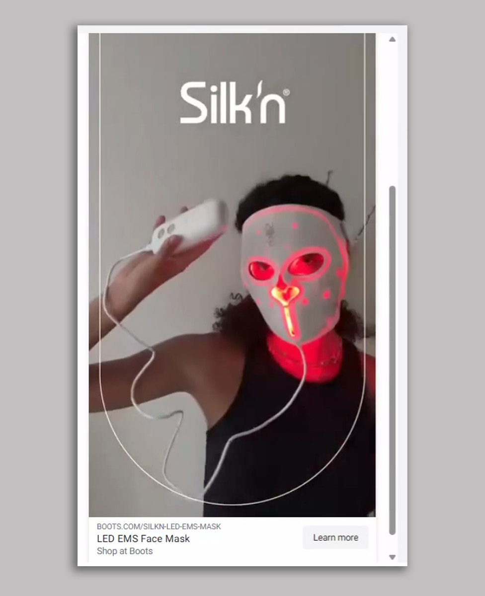 A Silk'n advert showing a woman in a black tank top with dark curly hair wearing a white LED face mask and holding the controller while it beams red light on to her face