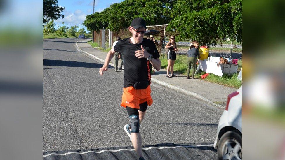 Karl Thompson running along a road in sunny conditions, wearing a black t-shirt and orange running shorts.