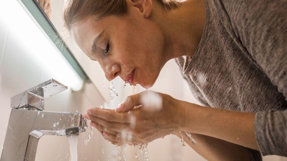 A woman with blonde hair and a grey top leans over a bathroom tap and splashes water over her face.