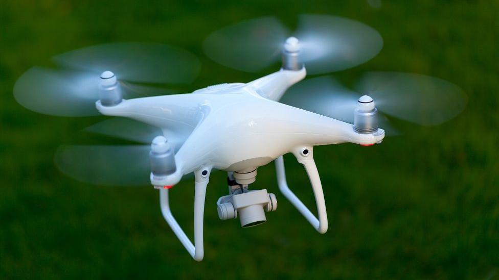 Flying drone with a camera taking pictures from a birds eye view. It is hovering above green grass to take aerial pictures.