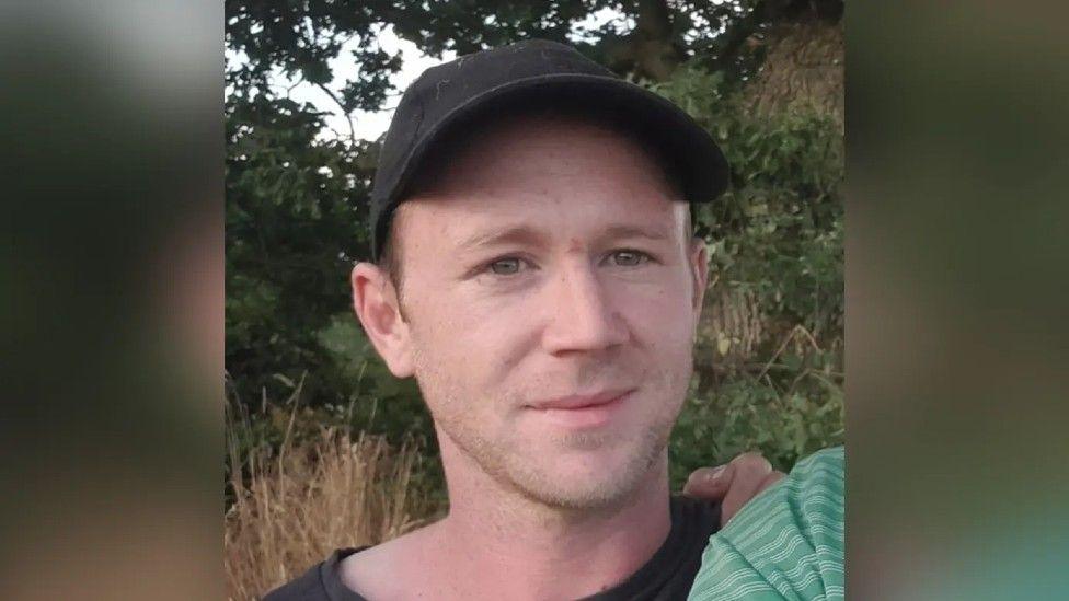 Michael O'Dwyer appears wearing a black baseball cap. He is fair haired and is unshaven. He is standing outside. There are some bushes and trees behind him.