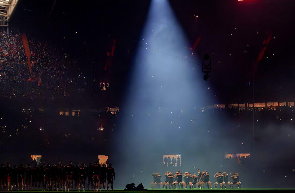 The All Blacks perform a synchronised haka under a dramatic spotlight on a dimly lit stadium field.