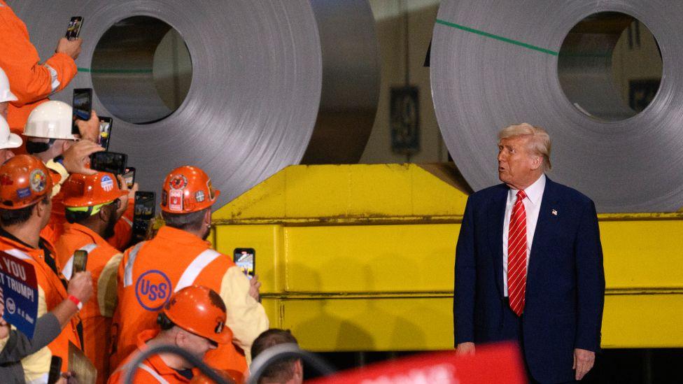 Donald Trump speaking to supporters at a steel factory. He is wearing a dark suit, a white shirt and red tie as men wearing hi-vis jackets and hard hats take pictures of him