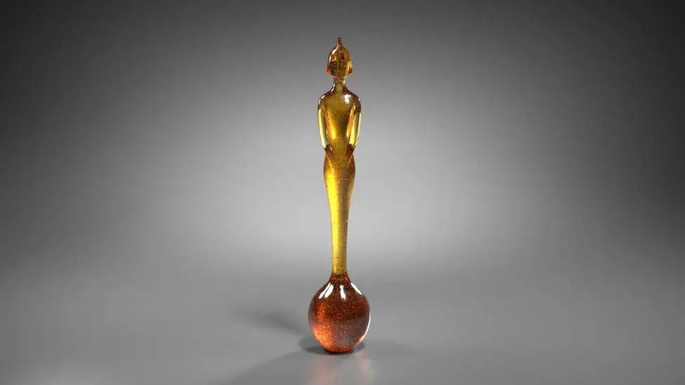 A tall, thin trophy made from amber resin stands against a grey background. It's in the shape of a human figure and stands on a round base that looks like a drip of honey.