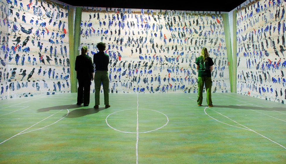 Three people standing in a room with a football pitch projected onto the floor and Lowry figures on the walls