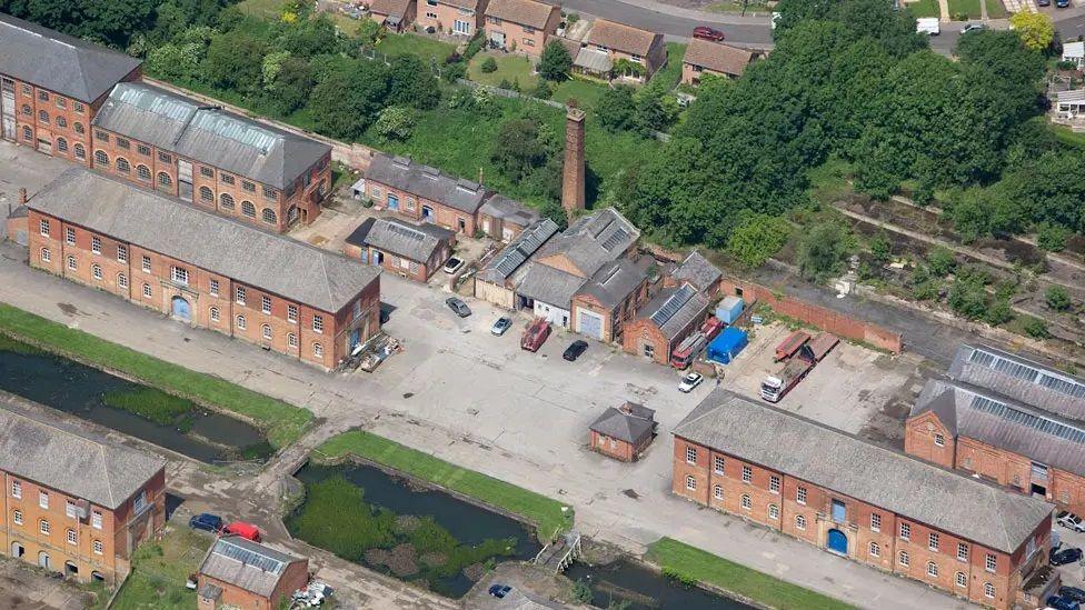 Aerial shot of The Depot, showing long narrow two-storey brick buildings. One has a tall chimney. There are some cars parked between the buildings. A canal with a grass verge can be seen towards the bottom of the picture. There are trees separating The Depot from a residential street at the top of the image.