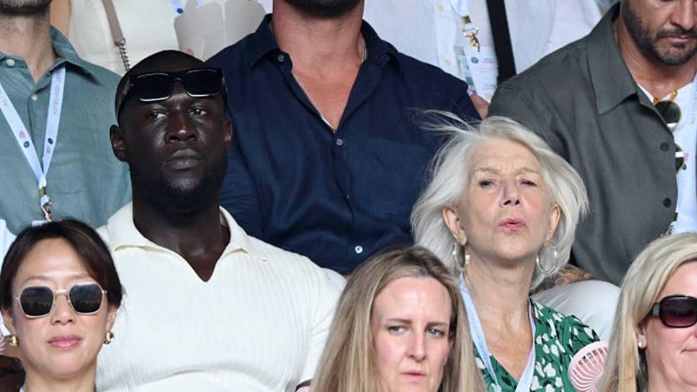 Stormzy, wearing a white polo shirt, sits next to Helen Mirren and Taylor Hackford. Mirren and Hackford have expressions of slight shock on their face.