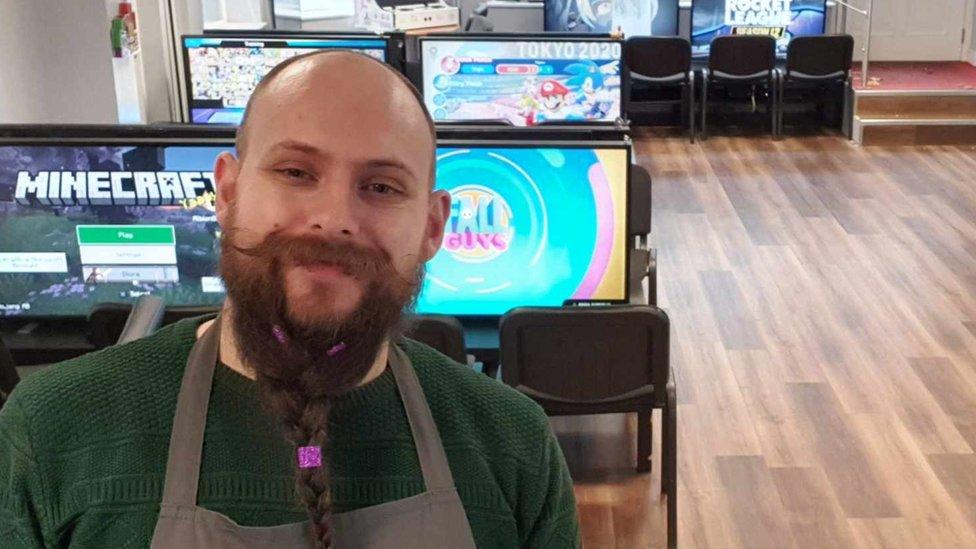 A man with a long beard, tied in a plait that hangs down to his chin, and a long moustache with the ends waxed into curls, stands in front of screens with video games playing on them. He's smiling and wearing a green sweater with a light grey apron over the top. The laminate wood floor behind him has six gaming stations set up on it, each one with two chairs and a large monitor. One is displaying the Minecraft title screen, another the Fall Guys title, one is showing Mario and Sonic at the Olympic games and the other is displaying the Rocket League title screen. The other two are obscured.