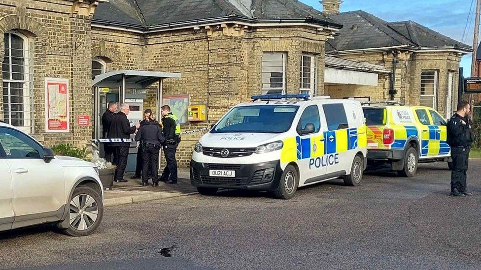 Two police cars parked outside the Harwich Town railway station. There is a police cordon and a group of people standing behind the police cars. One male officer is standing in front.