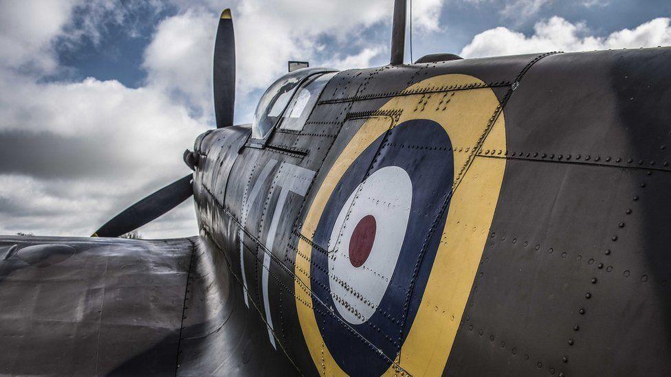 A close-up view of a the World War Two aeroplane the Spitfire, showing the left side painted grey. There is a yellow, blue, white and maroon circular log on its side above its wing. Beyond is the clear glass/plastic of the cockpits and its propeller. 