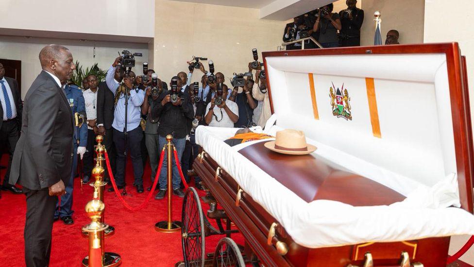 Kenya's President William Ruto stands before Raila Odinga's open casket which stills on a carriage whiles and his fedora cat placed centrally on the wooden coffin. A bank of photographers  capture the moment.