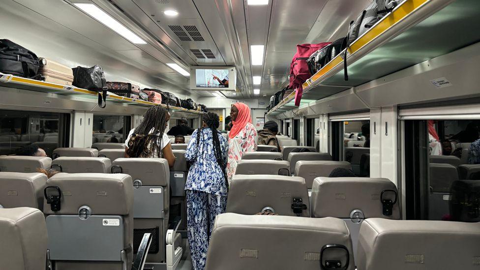 Interior of the train showing three seats on one side and two seats on the other. They are all light grey.