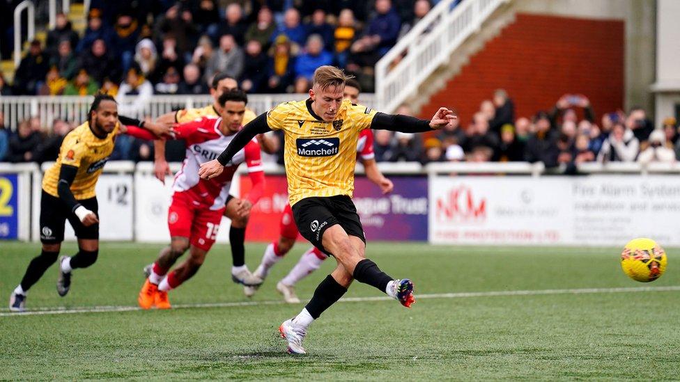 Maistone United player Sam Corne wears a yellow and black football kit and kicks a yellow ball on a pitch with fellow players behind