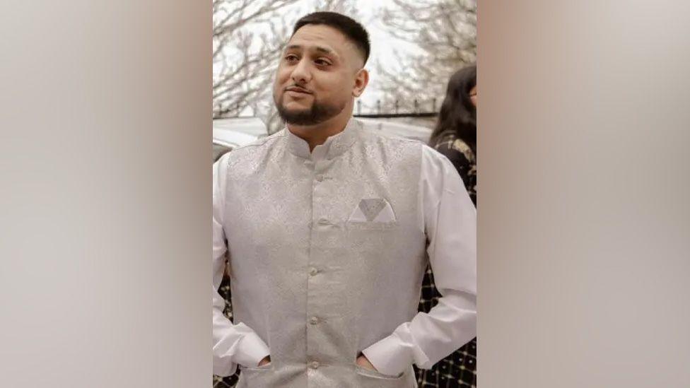 A photograph of Mohammed Duraab Khan wearing a white shirt and silver waistcoat and he is stood with his hands in his pockets