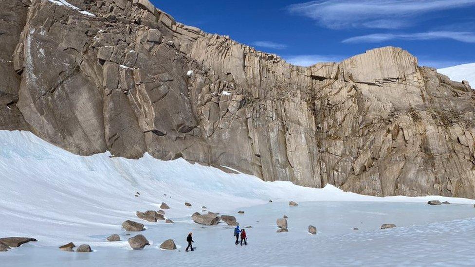 Large rocky cliff, snow on the ground, team walking in snow gear