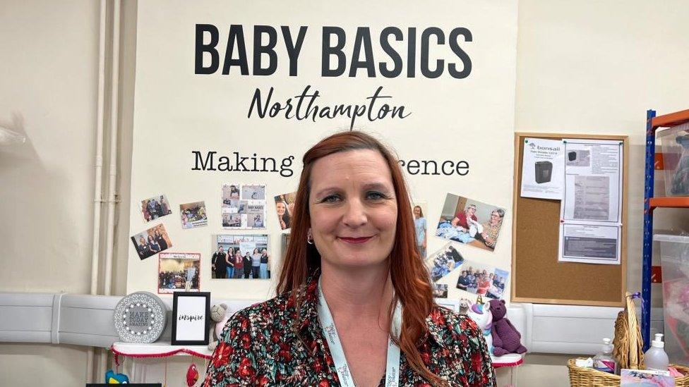 Woman with long brown/red hair wearing a lanyard in front of a Baby Basics sign