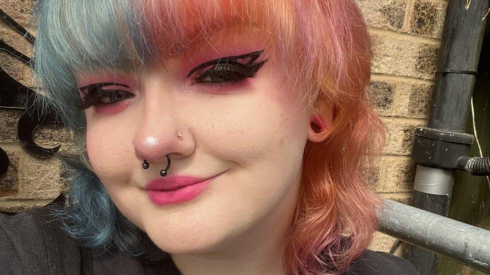 A selfie of Saryna Glazebrook, she has half blue and half pink hair, and is smiling at the camera. The background is of a brick wall.