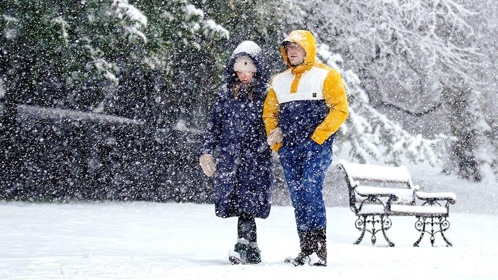 Two people walk in the snow in Buxton, Peak District with a bench and trees in the background, on 8 February