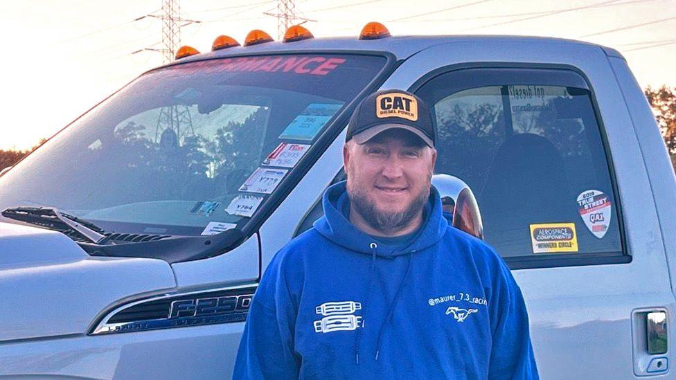 A half length portrait of Ben in front of a cab of a pick up truck. It is evening and he is in a blue hoodie and is wearing a baseball cap with a CAT company logo