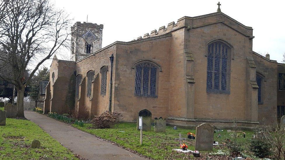 Exterior of the 1,000-year-old St Cuthbert's Church which has stained glass windows. There is a green garden with a graveyard in front.