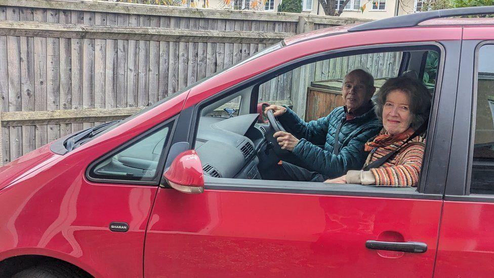 Richard and Helen Dodd sitting in the front seats of their red people carrier. Mr Dodd in  a blue padded anorak is at the wheel and Mrs Dodd in a striped jumper is resting her arm on the open passenger window 