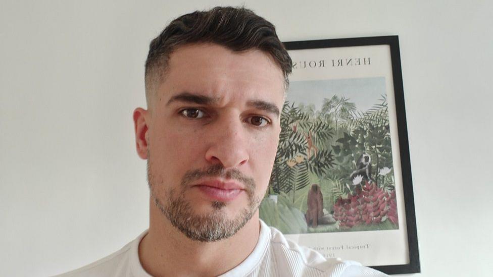 Stefan Bratu standing in front of a white wall in his home and a painting of a forest/jungle scene. He is wearing a white top and has short brown hair and a beard. He has a serious expression on his face.