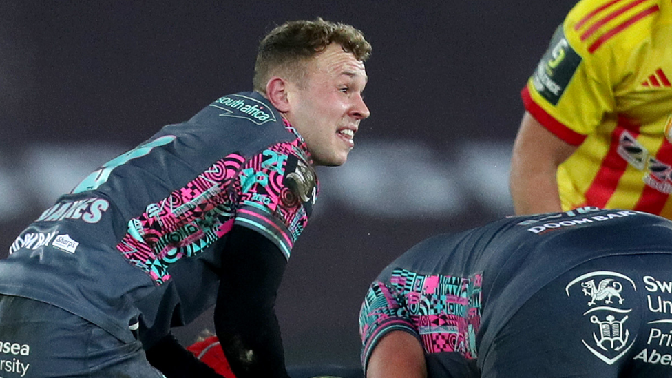 Luke Davies on the pitch during a rugby match. He is looking up to pass the ball from the ruck.