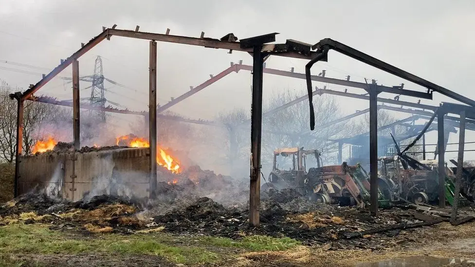 The aftermath of the fire, including hay burning on the left hand side and three wrecked tractors, with just the barn's metal frame left after the blaze