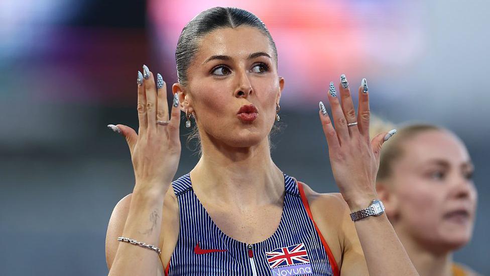 Close-up photo of athlete Amy Hunt. Her lips are pursed together and she has her dark hair slicked back. She has an earring in each ear and has both her hands held up near to face to show off her nail art which is white nails with silver 3D balls on them. She also has a ring on one finger of each hand. She is wearing a navy blue and white striped Team GB vest and also has jewellery on both of her wrists.