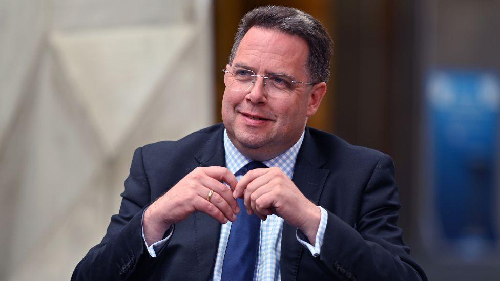 Craig Hoy, a man with short dark hair and glasses, photographed in a medium close-up. His hands are touching in front of his chest. He is wearing a dark suit, chequered blue and white shirt and navy blue tie. 