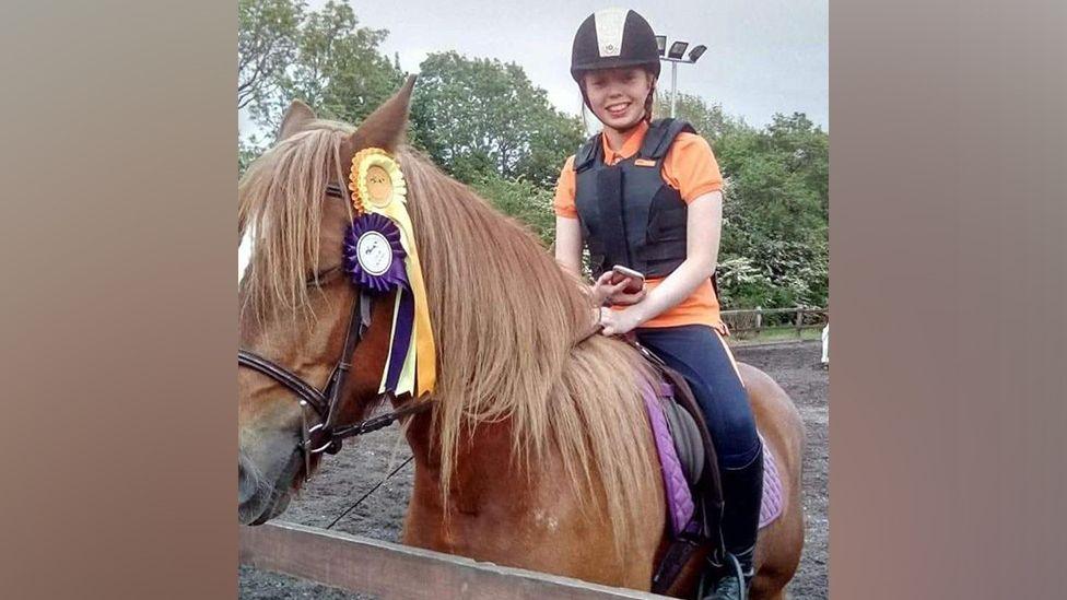 Caris Graham is sitting on a horse which has two rosettes hanging from its bridle. The horse is brown with a white patch along its nose. Ms Graham is holding a mobile phone in one hand and the bridle with the other. She is wearing an orange t-shirt and a black protective vest and riding helmet.