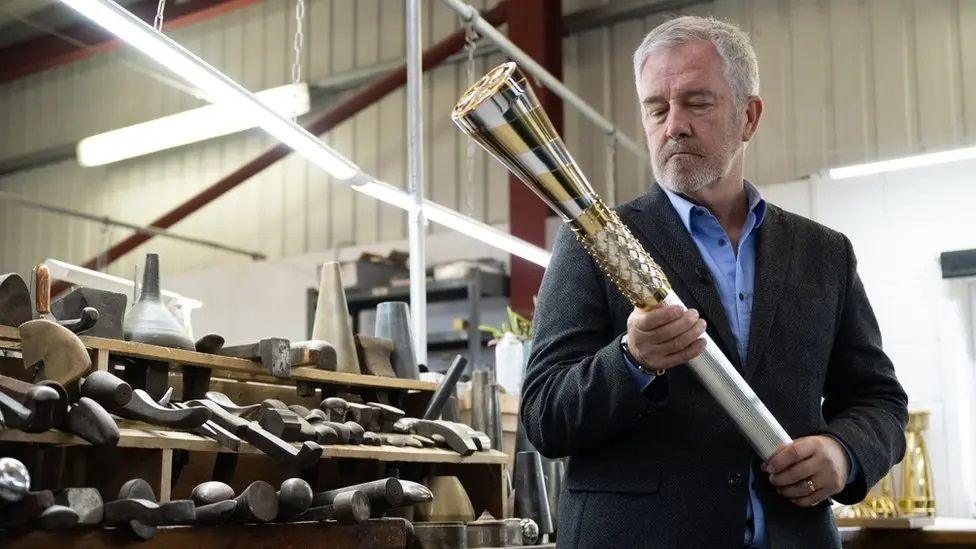 Mike McCarthy, a man with short, white hair and a short, white beard. He's holding the "Baton of Hope", which is silver and gold.