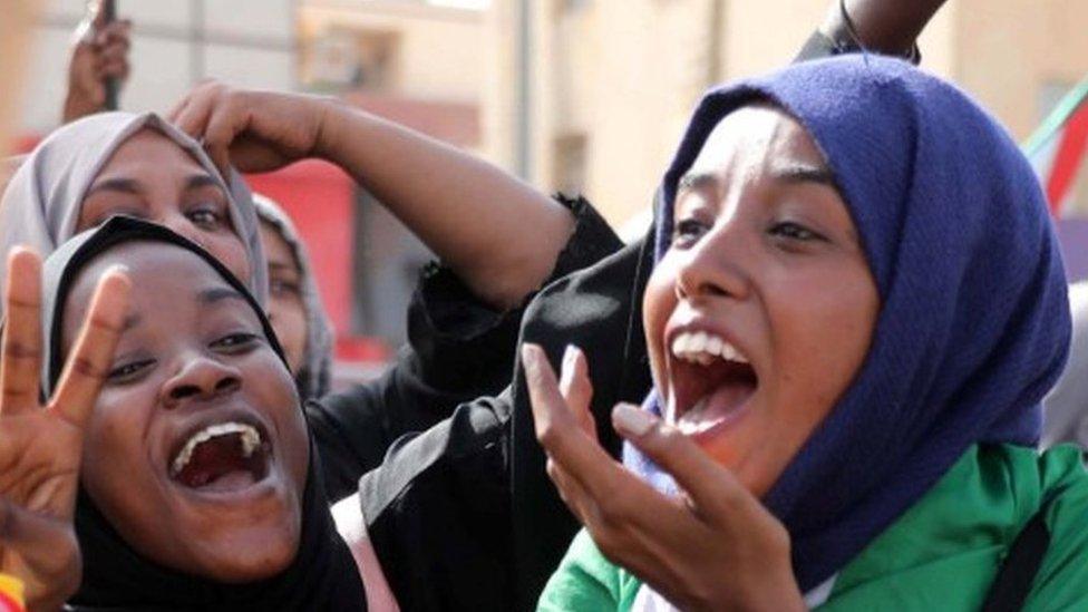 Young, female protesters react to news of the power-share deal with visible excitement, smiling, shouting and gesturing with their hands.