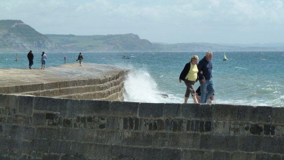 Five people walk along The Cobb as a wave sends up a high plume of spray. The curved breakwater is built from mortared stone