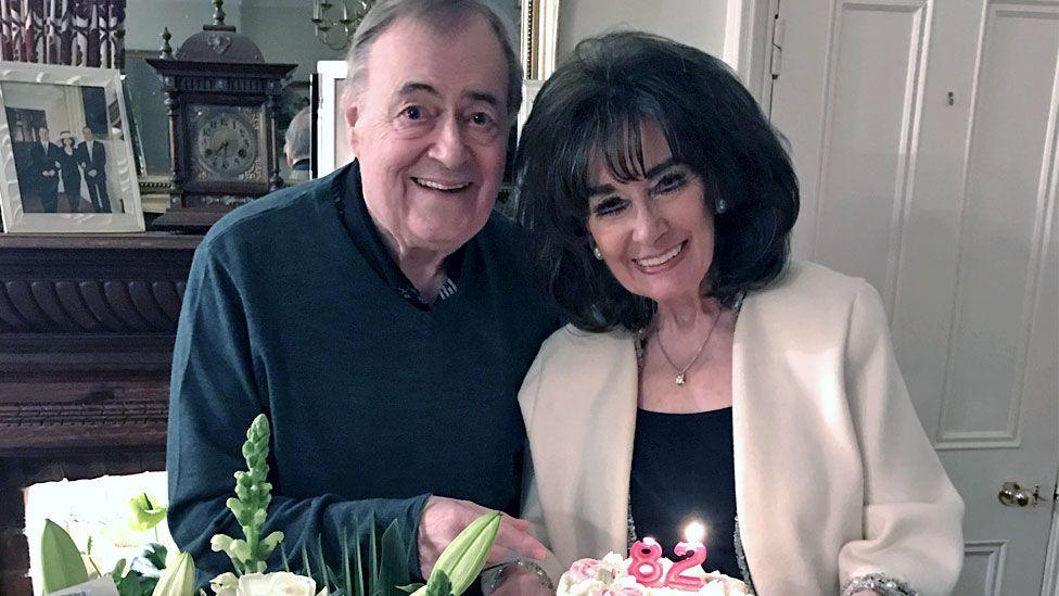 Prescott next to his wife, Pauline. Both are smiling and she is holding a birthday cake with two candles with the numbers 8 and 2