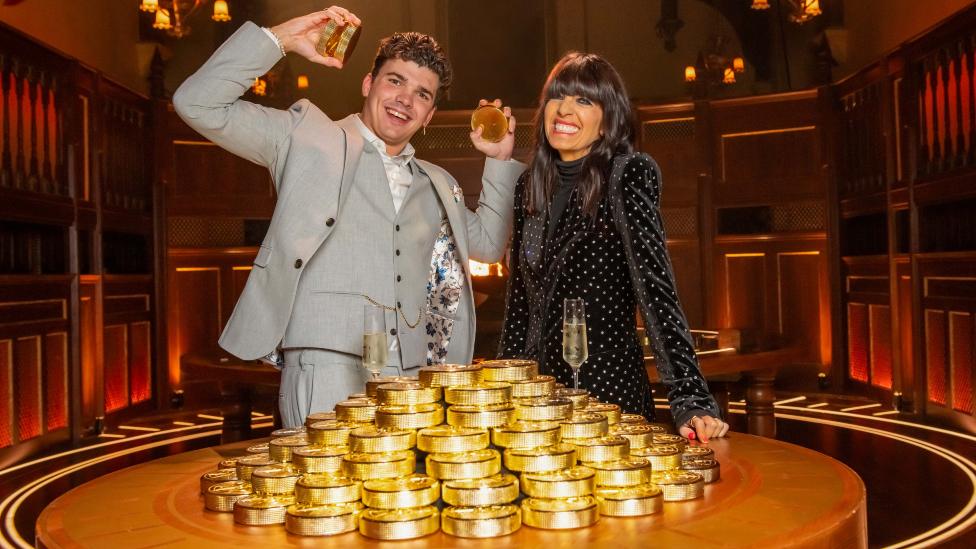 The Traitors winner Harry Clark with presenter Claudia Winkleman holding gold discs, standing by a table piled up with more gold discs