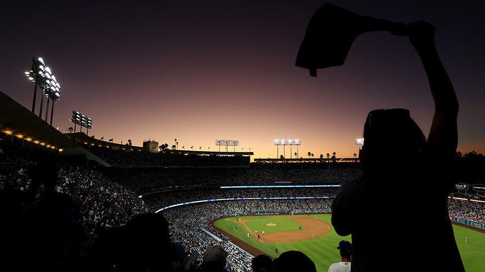 A packed baseball stadium at sunset with bright floodlights illuminating the field. A silhouetted fan waves a towel in the foreground as players are positioned on the diamond.