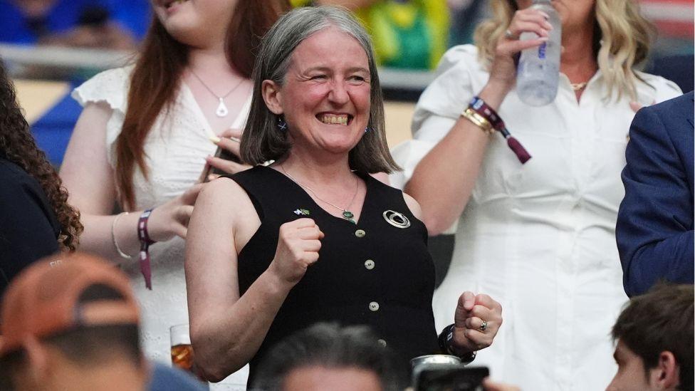 Maree Todd, who has shoulder-length grey hair, smiles with clenched fists in a football stadium stand 