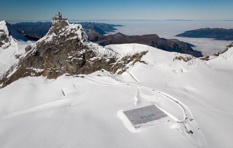 A zoomed-out aerial view shows the giant postcard on the Aletsch Glacier, with an expanse of snow all around it and the peaks rising behind