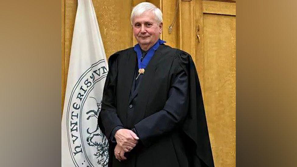 Steve McAdam is wearing a dark blue collar, a black jacket and a long blank gown. He is standing in front of a wooden doorway and white flag with blue lettering. 