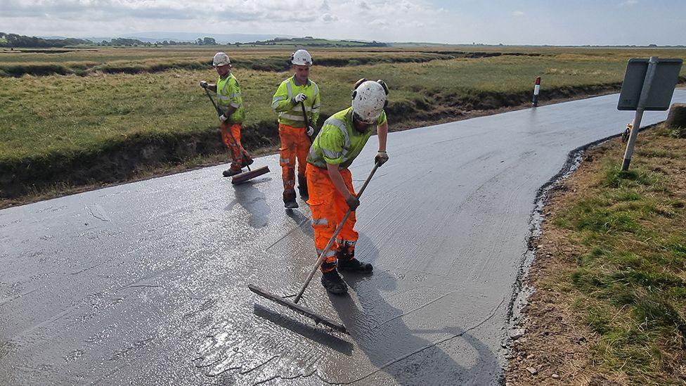 Three workers wearing high vis clothing and helmets, using long flat, rakes to smooth out the surface of the road