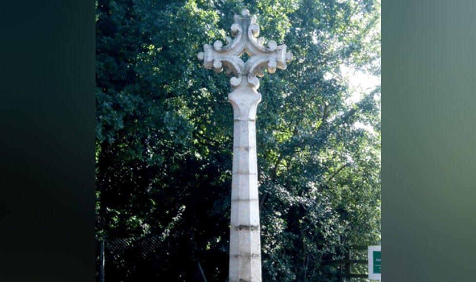 A tall, thin pale stone monument stands upright near some trees, and atop it is an ornate cross shape. 