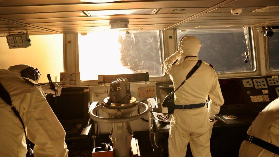 Two figures stand in a room of the HMS Diamond, looking out at a fiery scene