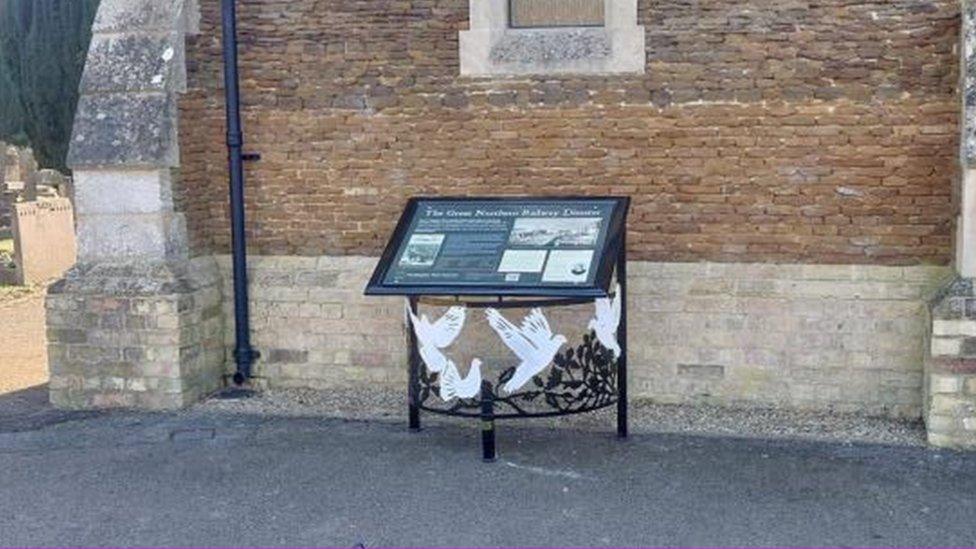 Black-framed interpretation board alongside stone wall of cemetery chapel