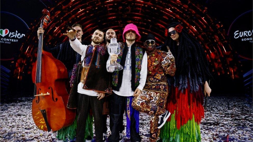 The six male members of Kalush Orchestra dressed in bright costumes holding the iconic glass microphone Eurovision trophy following their 2022 victory in Turin