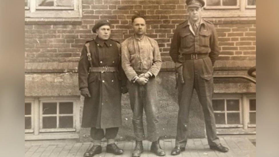 Black and white picture of Capt Cross with Höss in handcuffs and a guard standing by his side