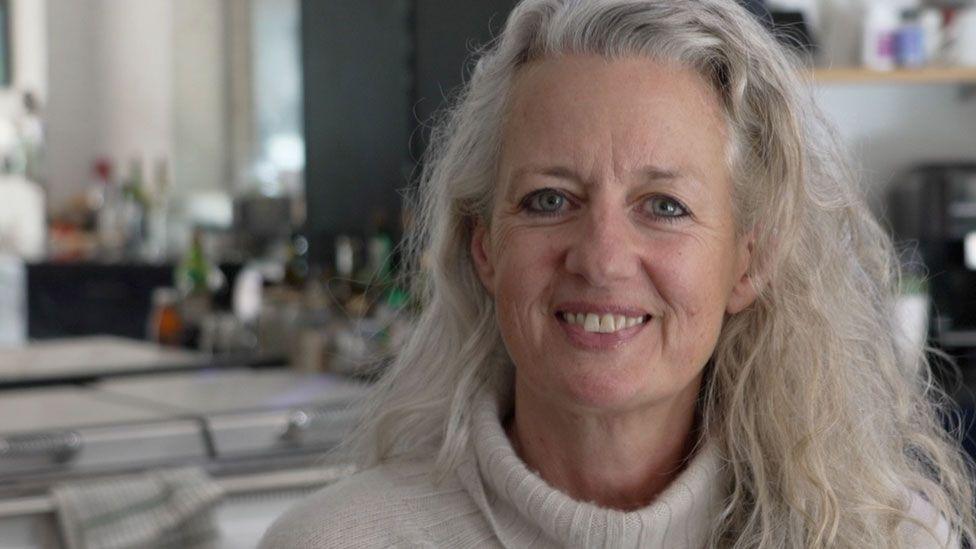 A close-up of Gay Longworth. She has long white hair and is wearing a beige polo-neck jumper. She is smiling and she is sitting in her kitchen with a white range behind her. 