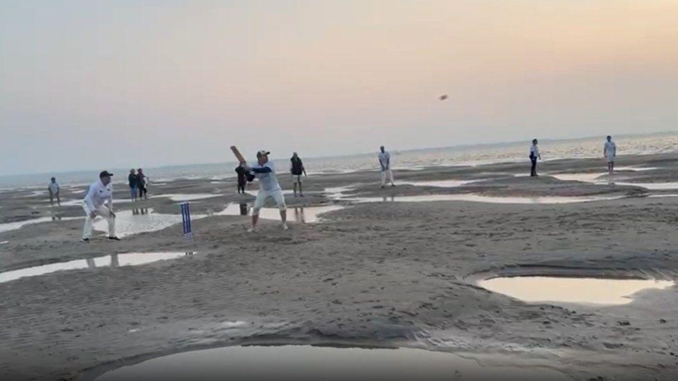 Sea and large puddles with sea beyond blue cricket stumps and players wearing cricket whites in action